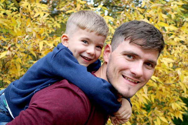 El padre fuerte sostiene a su pequeño hijo guapo lindo en su espalda al aire libre en el bosque — Foto de Stock