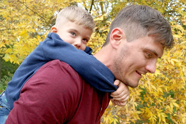 The strong father holds his cute handsome small son on his back outdoors in the woods — Stock Photo, Image