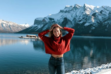 Young blonde with white smile and big lips. Red oversize jersey, flat belly, blue jeans. Raised hands up to the head. Powerful mountains and lake on the background. Cold winter weather. 