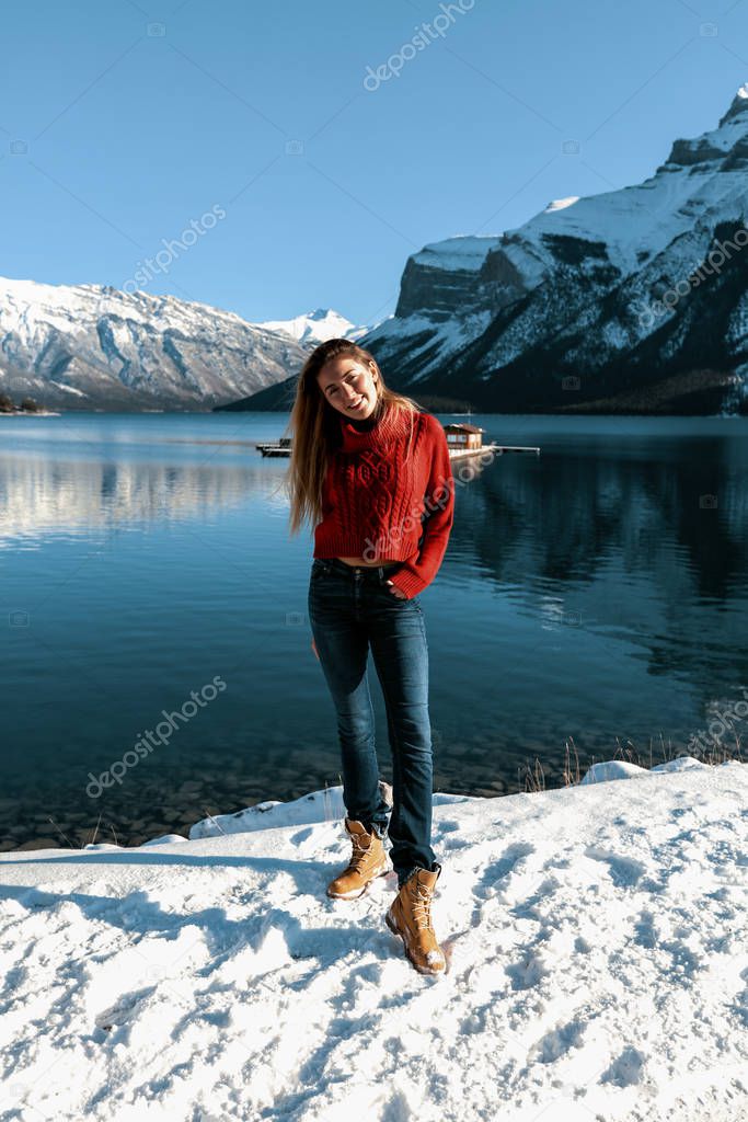 Funny lady with perfect slim body wearing blue jeans and red sweater. Long hair waving on the wind. Cold winter day outdoor. White snow, lake and mountains on the background. Smiling cheerful girl