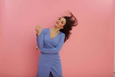 Tutkulu kız stüdyoda tek başına eğleniyor pembe arka planda duruyor ve uzun güzel saçlarını sallıyor. Şık bir makyaj ve göz alıcı bir elbise. Büyük dudaklar ve güzel gözler..