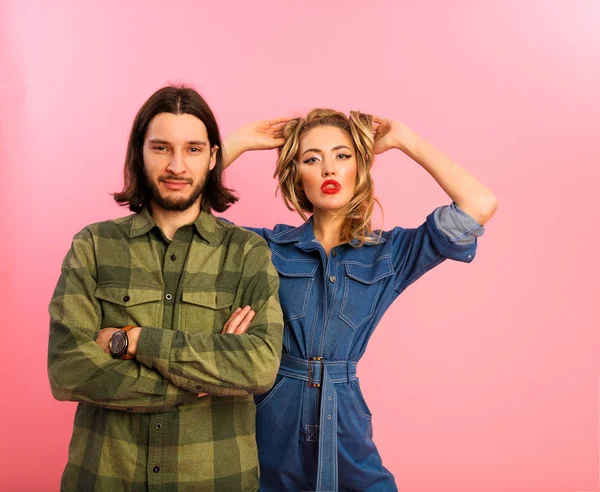 Amazing young lady with red big lips standing behind the modern guy in green shirt. Her arms on the head, holding long volume hair. He has dense beard and brunette long hairstyle. Pink background