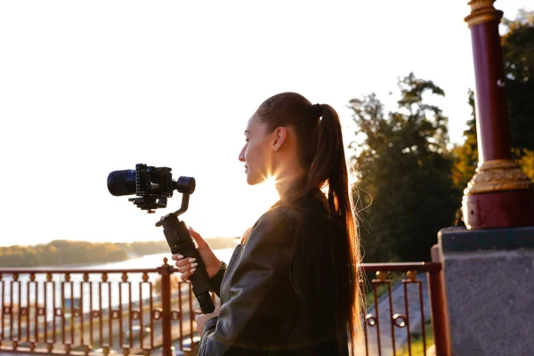 Attractive young brunette holding camera stabilizer and making a movie outdoor in the morning. Sun rising up above the river, summer warm weather and green trees. Wearing black leather jacket.