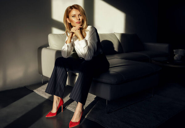 Элегантная женщина, сидящая одна на диване в комнате. Классический костюм с красной яркой обувью на высоких каблуках. Положите локти на колени, наклоните голову. Блондинка объем волос и голый макияж на ее лице
