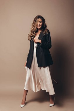 Yüksek topuklu ayakkabılı genç model stüdyoda duruyor. Zarif, modern bir elbise ve siyah bir ceket. Şık kıvırcık saçlar ve doğal makyaj. Kahverengi arka plan. Uzun ve zayıf kız.