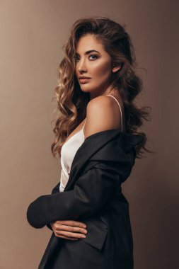 Ciltli kıvırcık saçlı ve modern çıplak makyajlı güzel bir kız stüdyoda duruyor. İnce vücudunda siyah ceket ve beyaz ipek elbise var. Yumuşak omzunu gösteriyor. Kahverengi arkaplan 