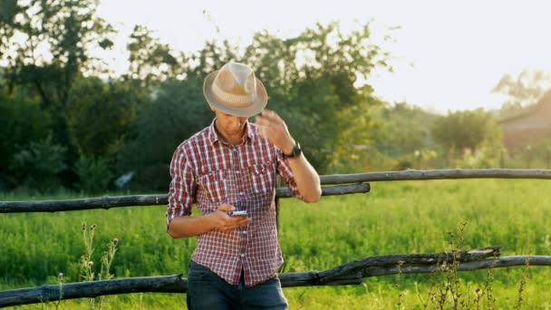 Joven con sombrero usa Smartphone. Zona rural. — Vídeo de stock