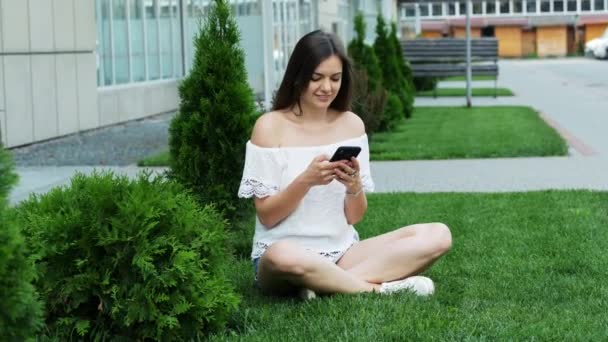 Junge schöne Frau benutzt Smartphone, während sie auf dem Gras in der Nähe des Hotels sitzt — Stockvideo