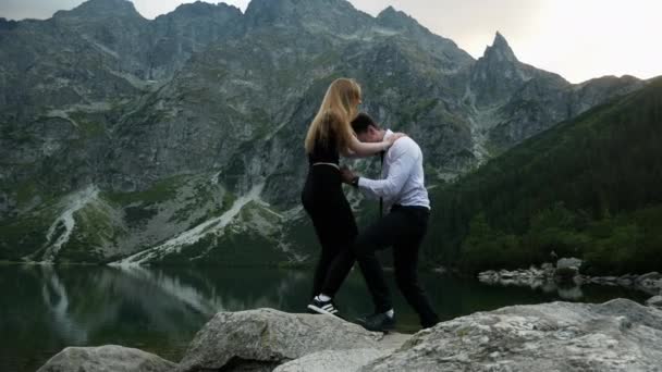 Мужчина в костюме опускается на колени и делает предложение своей любимой женщине возле горного озера, надевает обручальное кольцо — стоковое видео