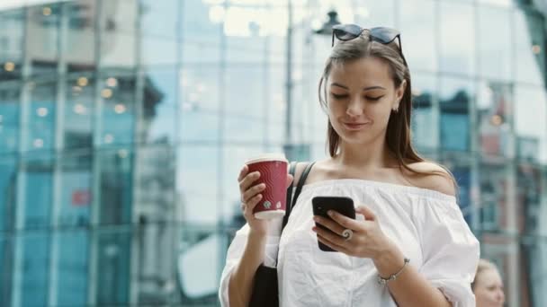 Lächelnde süße Frau mit einer Tasse Kaffee nutzt Handy auf Geschäftsstraße — Stockvideo