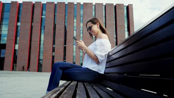 Gelukkig vrouw in een zonnebril gebruik maken van mobiele telefoon tijdens het zitten op de bank op straat, moderne bakstenen gebouw — Stockvideo