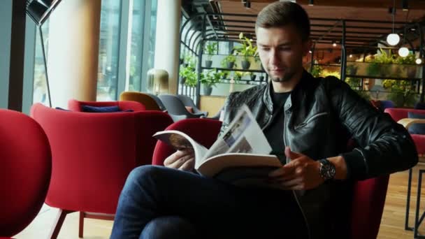 Homem turístico atraente lê uma revista enquanto espera em um hotel moderno — Vídeo de Stock
