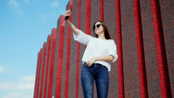 Vacker kvinna använder en mobiltelefon för selfie nära en modern tegelbyggnad — Stockvideo
