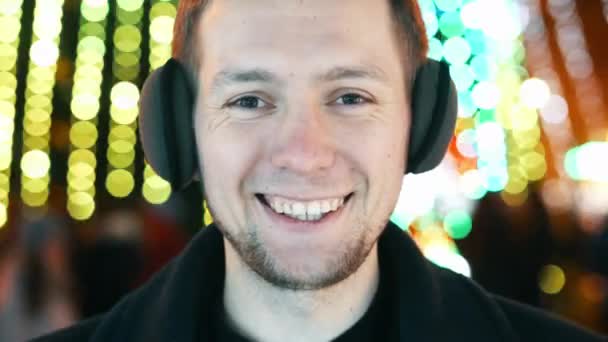 Fiatal vidám férfi portré meleg fejhallgató mosollyal és a kamerába nézve, bokeh karácsonyfüzér fények — Stock videók
