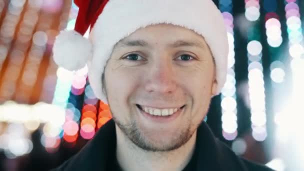 Jeune homme gai portrait avec le sourire chapeau de Nouvel An et en regardant la caméra, bokeh de lumières guirlande de Noël — Video