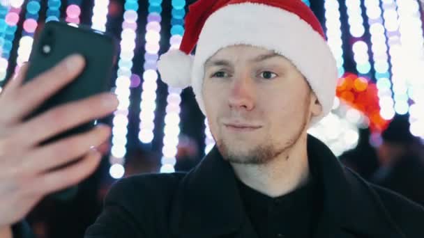 Vrolijke man neemt selfie en bouwt grappige gezichten op mobiele telefoon, gekleed in kerstmuts. Kerstboom — Stockvideo