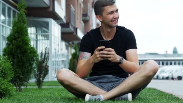 Attraktiv man använda mobiltelefon när du sitter på gräset nära hotellet, chatta med vänner — Stockvideo