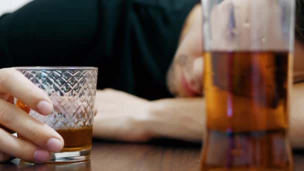 Ung berusad man dricker whisky ur ett glas och somnar på bordet, en halvtom flaska whisky i förgrunden — Stockvideo