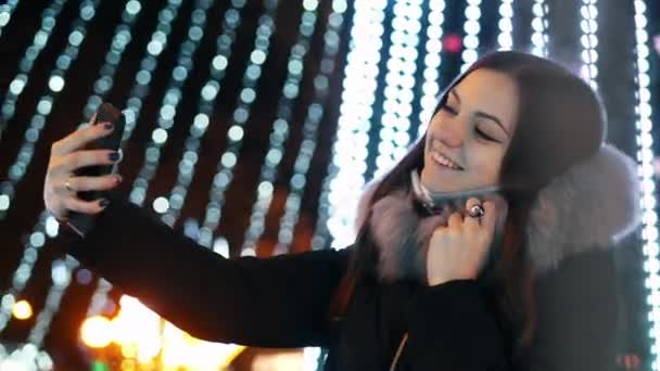 Genç ve güzel bir kadın, yılbaşı ışıklarıyla süslenmiş bir şekilde cep telefonuyla selfie çekiyor. — Stok video