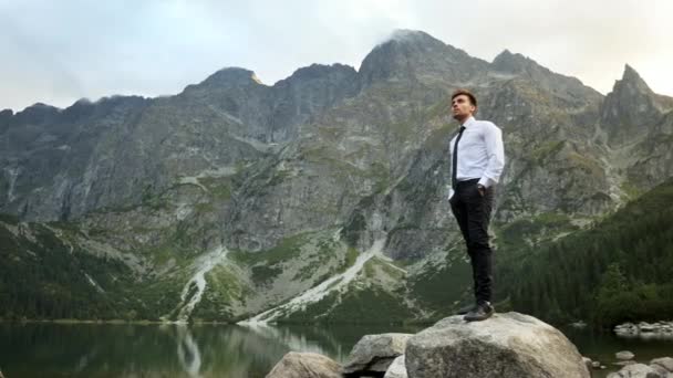 Successful confident businessman standing on the rock near a lake in the mountains at sunset, looks at the watch on his hand — 비디오