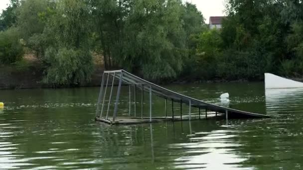 Hombre extremo en un traje con corbata y gafas de sol monta en wakeboard y saltando en un trampolín en un lago — Vídeo de stock