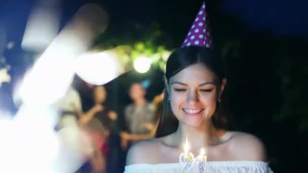 Mulher bonita fica com um bolo e velas para o seu aniversário, faz um desejo, amigos estão dançando no fundo, festa à noite — Vídeo de Stock