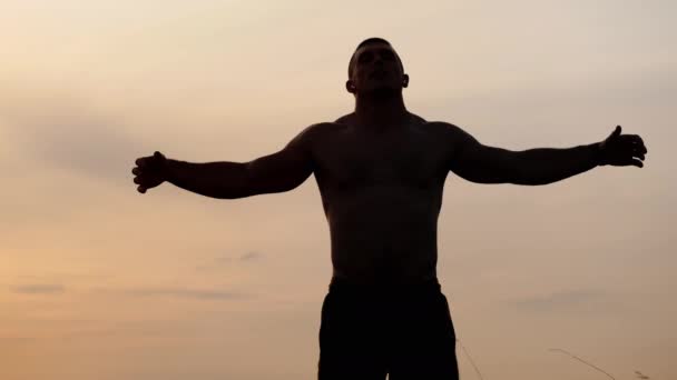 Silhouet van een sporter met een kale romp poseert toont zijn spieren en biceps tegen de hemel bij zonsondergang. Sport, gezondheid en fitness — Stockvideo