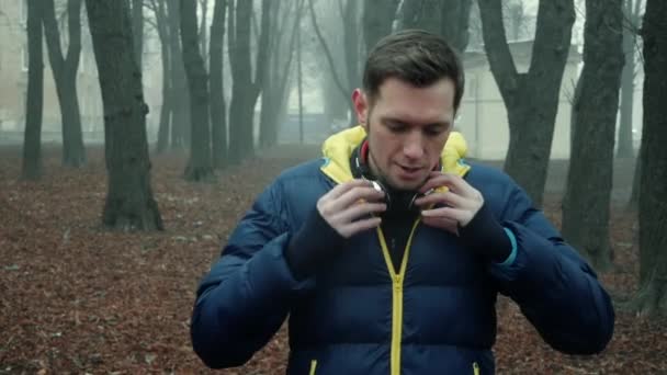 男性アスリートは、都市公園、冷たい霧の朝にワイヤレスヘッドフォンを置きます — ストック動画