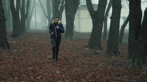 Чоловік-спортсмен бігає в міському парку на холодний ранок в тумані, знімає свої бездротові навушники — стокове відео