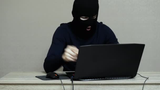 Смішний чоловічий хакер в чорній масці Балаклава роблять хакерську атаку на ноутбук, дивлячись на камеру — стокове відео