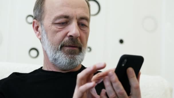 Man van middelbare leeftijd met een grijze baard gebruikt de telefoon thuis, close-up — Stockvideo