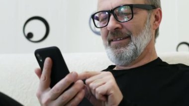 Middle-aged hipster man with a gray beard and glasses use mobile phone at home in close-up