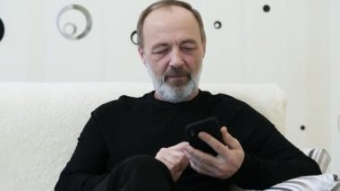Gri sakallı orta yaşlı bir adam telefonunu evde kullanıyor, yakın plan.