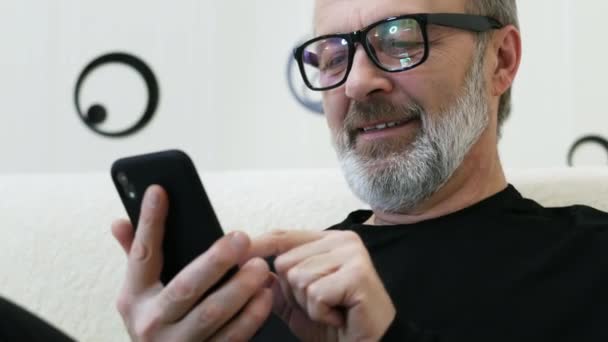 Middle-aged hipster man with a gray beard and glasses use mobile phone at home in close-up — 비디오
