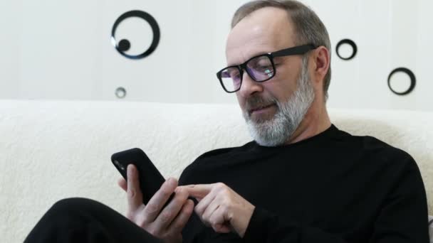 Middle-aged hipster man with a gray beard and glasses use mobile phone at home — 비디오