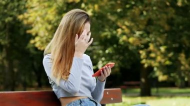 Cute redhead young woman using smart phone on a bench in a park, young woman smiling while surfing internet