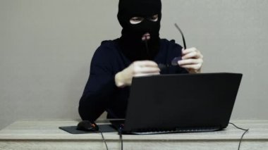 Siyah maskeli bir erkek bilgisayar korsanı gözlük takar ve dizüstü bilgisayara hacker saldırısı yapar.