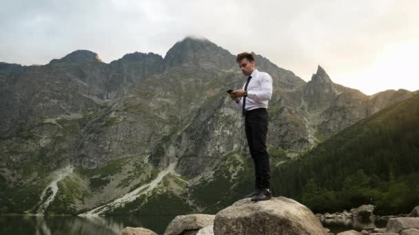 Porträt eines erfolgreichen selbstbewussten Geschäftsmannes, der bei Sonnenuntergang sein Handy in der Nähe eines Sees in den felsigen Bergen benutzt — Stockvideo