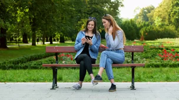 Dos mujeres violan la cuarentena y se sientan en un banco en el parque y usan el teléfono móvil — Vídeo de stock