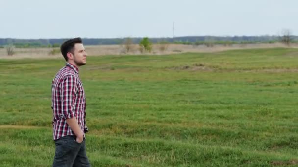 Chico hipster feliz camina en el campo al atardecer, sonríe, se siente libre . — Vídeo de stock