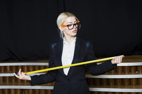 Hermosa mujer de negocios rubia con cinta métrica en la oficina loft en gafas naranjas y traje negro. Concepto empresarial . — Foto de Stock