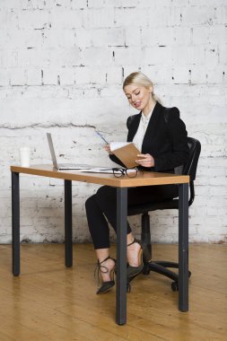 Genç sarışın güzel işkadını dizüstü bilgisayar, dizüstü bilgisayar ve gözlük kıyafetli bir office masada oturuyor. İş kavramı.