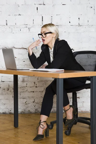 Νεαρή ξανθιά ομορφιά επιχειρηματίας που κάθεται σε ένα τραπέζι γραφείου με laptop, σημειωματάριο και γυαλιά στο κοστούμι. Επιχειρηματική ιδέα. — Φωτογραφία Αρχείου