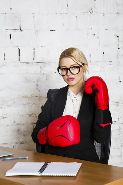 Νεαρή ξανθιά ομορφιά επιχειρηματίας που κάθεται σε ένα τραπέζι γραφείου με laptop, σημειωματάριο και γυαλιά σε κοστούμι και γάντια του μποξ. — Φωτογραφία Αρχείου