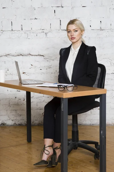 Genç sarışın güzel işkadını dizüstü bilgisayar, dizüstü bilgisayar ve gözlük kıyafetli bir office masada oturuyor. İş kavramı.