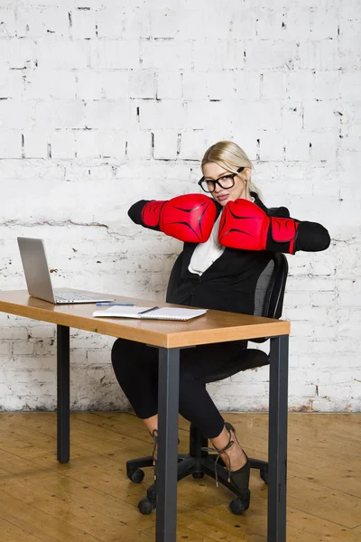 年轻的金发美女女实业家坐在办公室的桌子上, 笔记本电脑, 笔记本和眼镜的西装和拳击手套. — 图库照片