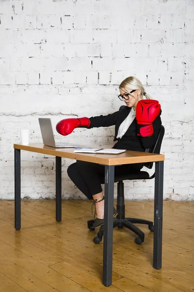 Young blond beauty businesswoman sitting at a office table with laptop, notebook and glasses in suit and boxing gloves. — Stock Photo, Image