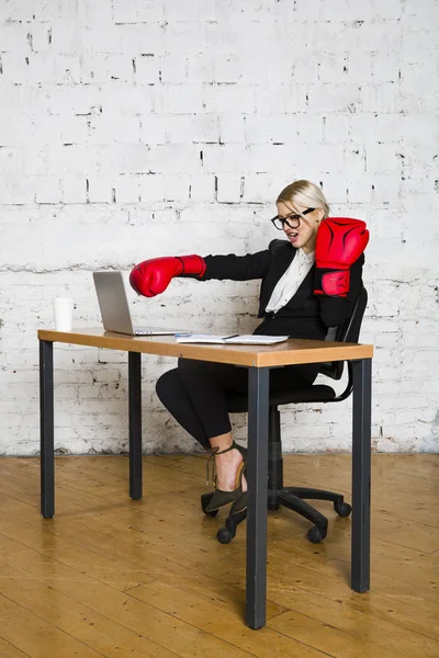 Young blond beauty businesswoman sitting at a office table with laptop, notebook and glasses in suit and boxing gloves. — Stock Photo, Image