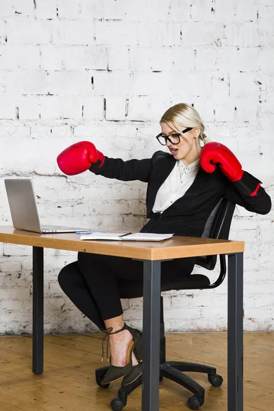 Young blond beauty businesswoman sitting at a office table with laptop, notebook and glasses in suit and boxing gloves. — Stock Photo, Image