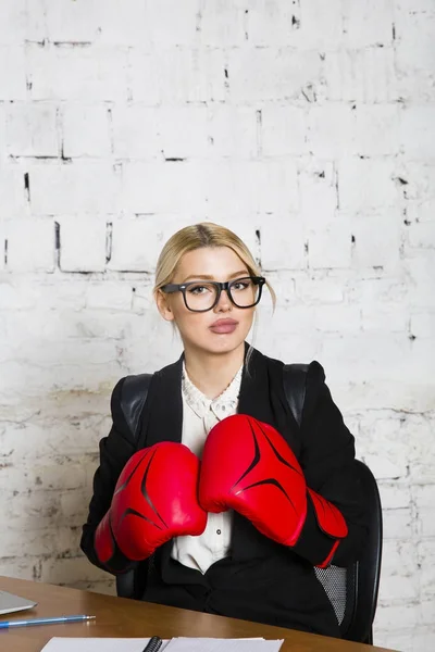 Junge blonde Schönheitsunternehmerin sitzt an einem Bürotisch mit Laptop, Notizbuch und Brille in Anzug und Boxhandschuhen. — Stockfoto